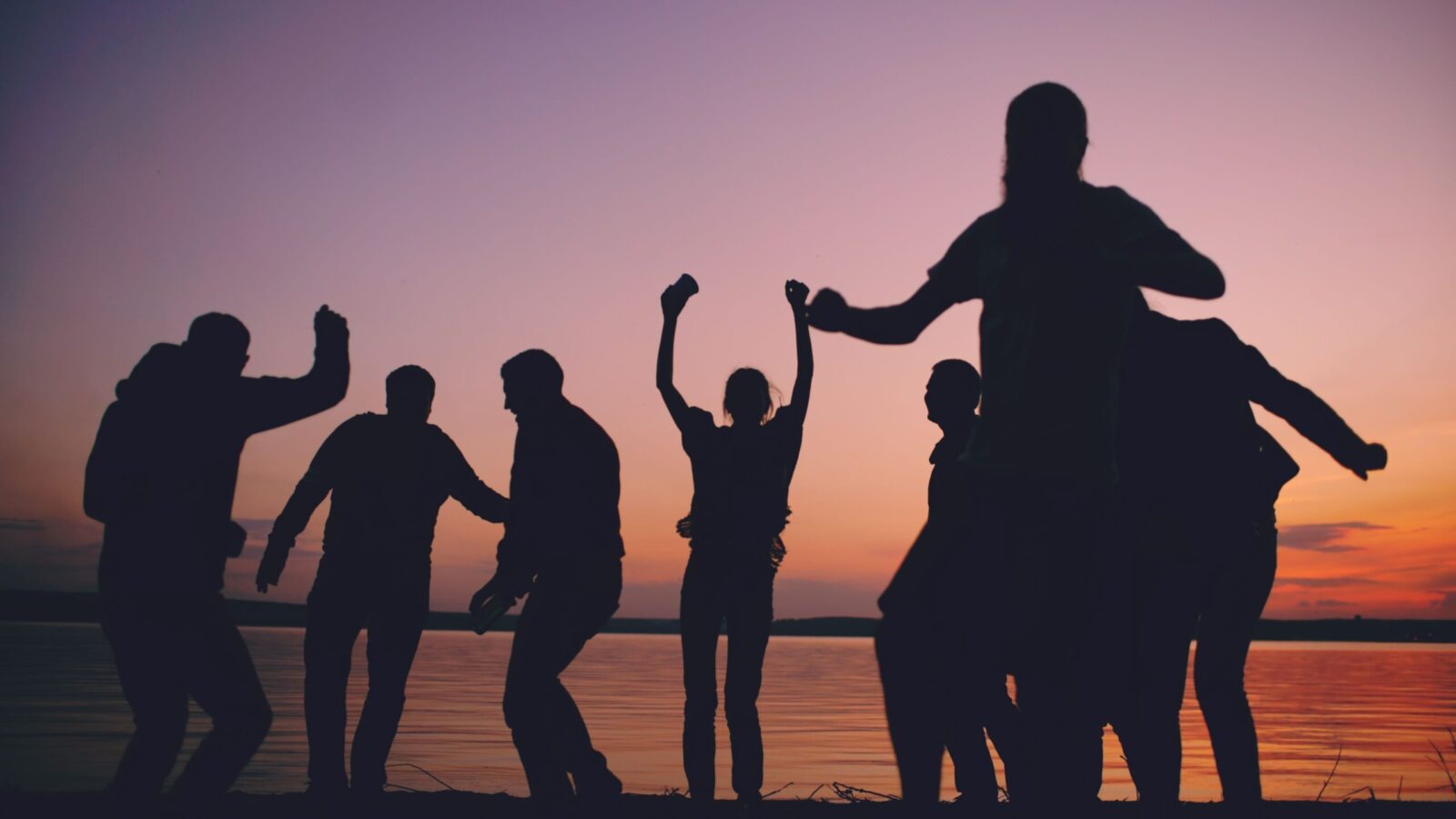 Group of joyful people dancing by a lake during sunset, creating a lively silhouette.